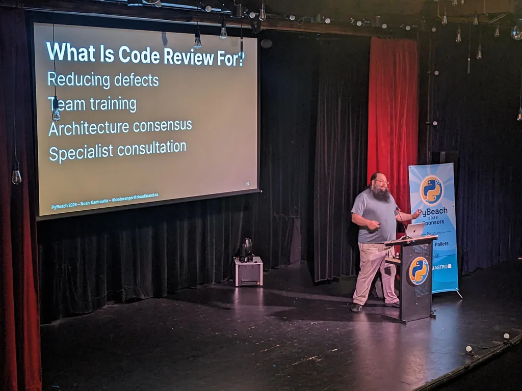 Noah Kantrowitz on stage with his 'What Is Code Review For?' slide: reducing defects, team training, architecture consensus, specialist consultation
